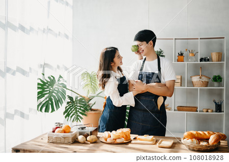 A man in the kitchen preparing a meal with Bread and fruit in cozy kitchen while his wife hugging him. sweet home. A man in the kitchen preparing a meal with Bread and fruit in cozy kitchen while his wife hugging him. sweet home. 108018928