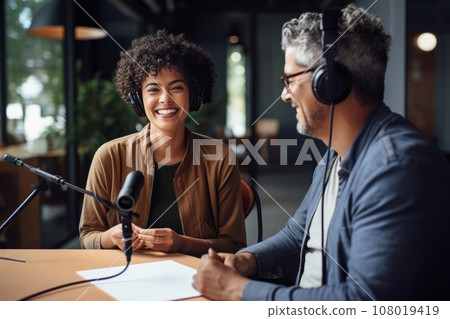 Beautiful multiethnic blogger couple hosting a podcast in a studio. Smiling woman and man wearing headphones sitting at a table with microphones and chatting cheerfully. Modern media technologies. Beautiful multiethnic blogger couple hosting a podcast in a studio. Smiling woman and man wearing headphones sitting at a table with microphones and chatting cheerfully. Modern media technologies. 108019419