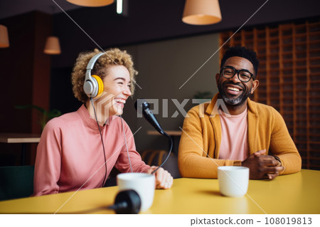 Beautiful multiethnic blogger couple hosting a podcast in a studio. Smiling woman and man wearing headphones sitting at a table with microphones and chatting cheerfully. Modern media technologies. Beautiful multiethnic blogger couple hosting a podcast in a studio. Smiling woman and man wearing headphones sitting at a table with microphones and chatting cheerfully. Modern media technologies. 108019813