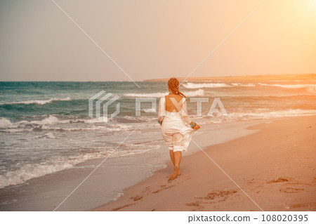 Model in boho style in a white long dress and silver jewelry on the beach. Her hair is braided, and there are many bracelets on her arms. 108020395