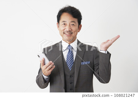 Middle-aged businessman holding a smartphone and pointing to the right side with his palm, white background 108021247