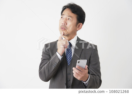 A middle-aged businessman holding a smartphone and thinking, white background 108021268