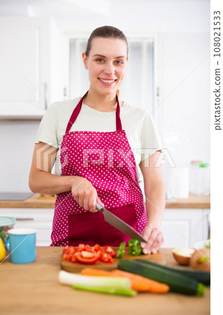 Positive young girl slicing vegetables for salad in cozy kitchen 108021533