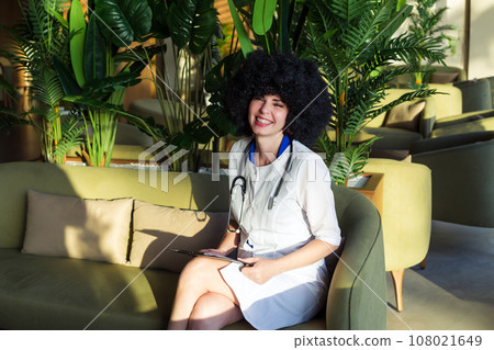 Nurse with black curly hair sits in hospital hallway, natural light 108021649
