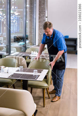 The owner of the cafe arranges chairs and tables before starting work 108021866