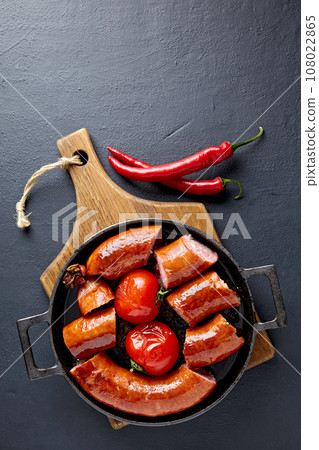 Sliced pork sausage fried in a pan with tomatoes in boiling oil on a wooden cutting board and hot peppers on a black concrete table Sliced pork sausage fried in a pan with tomatoes in boiling oil on a wooden cutting board and hot peppers on a black concrete table 108022865