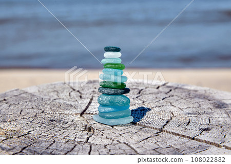 Balanced pyramid of sea shards of blue and green colored glass bottles on a wooden surface on the seashore. Sea-polished shards of glass bottles stacked on top of each other Balanced pyramid of sea shards of blue and green colored glass bottles on a wooden surface on the seashore. Sea-polished shards of glass bottles stacked on top of each other 108022882