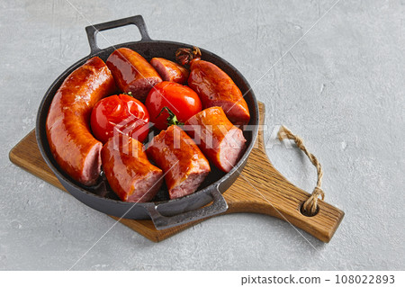 Fatty pork sausage cut into pieces and fried tomatoes on a black cast iron frying pan on a light concrete table 108022893