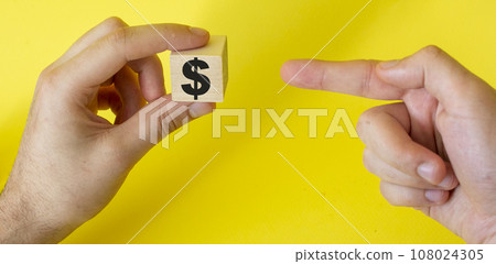 Hand holds a wooden cube with a dollar sign and points a finger at it on a yellow background Hand holds a wooden cube with a dollar sign and points a finger at it on a yellow background 108024305