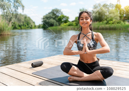 Young Indian woman practicing yoga meditation outdoors sitting in lotus position Padmasana on pier by the river. 108029525
