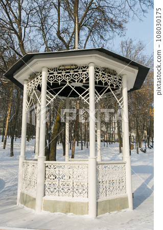 Beautiful white gazebo in a winter park against the backdrop of snowy trees. Park in city. 108030175