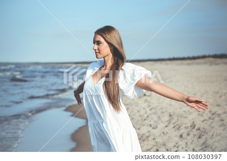 Happy smiling beautiful woman is on the ocean beach in a white summer dress, open arms Happy smiling beautiful woman is on the ocean beach in a white summer dress, open arms 108030397