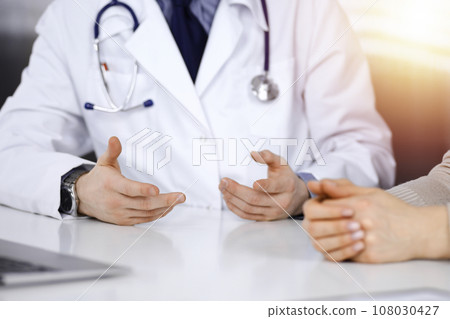 Unknown male doctor and patient woman discussing something while sittingin a darkened clinic, glare of light on the background. Close-up of hands 108030427
