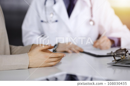 Unknown male doctor and patient woman discussing something while sittingin a darkened clinic, glare of light on the background. Close-up of hands Unknown male doctor and patient woman discussing something while sittingin a darkened clinic, glare of light on the background. Close-up of hands 108030435