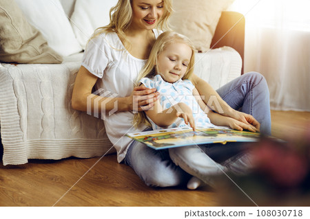 Happy family. Blonde young mother reading a book to her cute daughter while sitting at wooden floor in sunny room. Motherhood concept 108030718