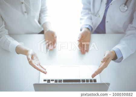 Doctor and patient sitting near each other at the table in clinic. The focus is on female hands pointing into tablet computer touchpad together. Medicine concept Doctor and patient sitting near each other at the table in clinic. The focus is on female hands pointing into tablet computer touchpad together. Medicine concept 108030882
