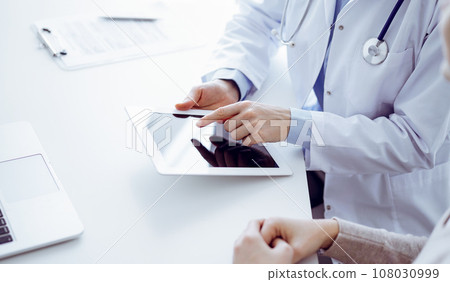Doctor and patient sitting at the table in clinic. The focus is on female physician's hands using tablet computer, close up. Medicine and healthcare concept Doctor and patient sitting at the table in clinic. The focus is on female physician's hands using tablet computer, close up. Medicine and healthcare concept 108030999