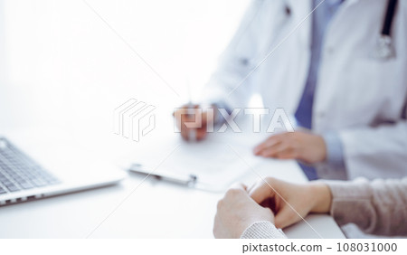 Doctor and patient sitting at the table in clinic. The focus is on female physician's hands filling up the medication history record form or checklist, close up. Medicine concept 108031000