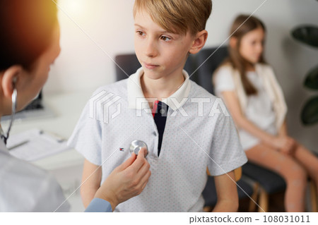 Doctor and kid patient are in the clinic. Physician in white coat examining a serious young boy with a stethoscope, close up. Medicine, therapy concept Doctor and kid patient are in the clinic. Physician in white coat examining a serious young boy with a stethoscope, close up. Medicine, therapy concept 108031011