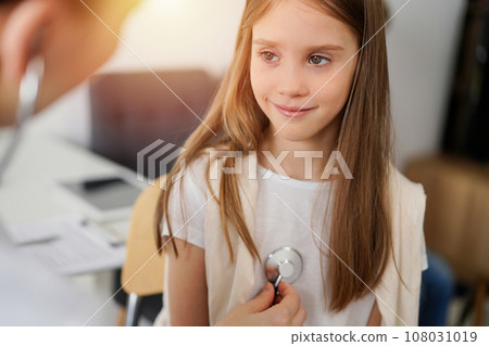 Doctor and kid patient are in the clinic. Physician in white coat examining a smiling young girl with a stethoscope, close up. Medicine, therapy concept 108031019