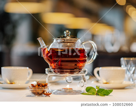 Glass teapot with warmer and candle, with hot herbal tea on table on restaurant background with bokeh. Tea concept with transparent glass teapot and cups on table in restaurant interior. 108032660
