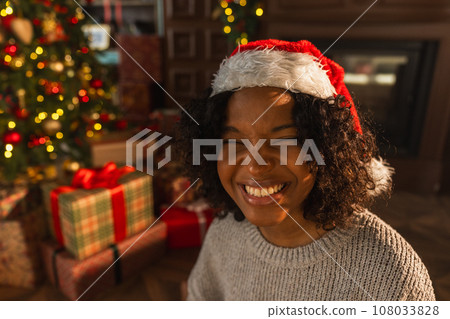 Merry Christmas. African American woman smiling near Christmas tree in classical dark interior. Happy girl in Santa hat in living room with fireplace Christmas tree gift boxes. Christmas eve at home 108033828