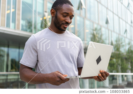 African American man freelancer using laptop typing chatting on urban street in city. Guy having virtual meeting online chat conference. Entrepreneur programmer working on laptop outdoors. Remote work 108033865