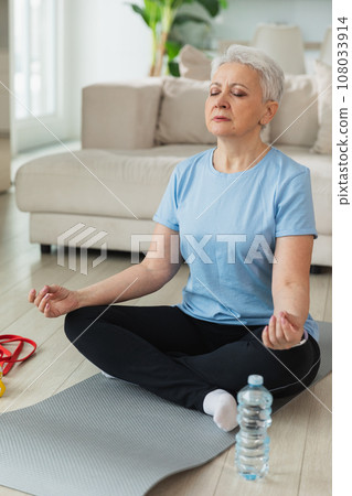 Yoga mindfulness meditation. Senior adult mature woman practicing yoga at home. Mid age old lady sitting in lotus pose on yoga mat meditating relaxing. Older middle aged woman doing breathing practice Yoga mindfulness meditation. Senior adult mature woman practicing yoga at home. Mid age old lady sitting in lotus pose on yoga mat meditating relaxing. Older middle aged woman doing breathing practice 108033914