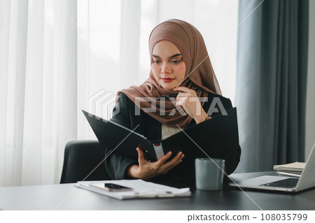 Young muslim millennial businesswoman in brown hijab headscarf working with financial reports while sitting in modern office. Young muslim millennial businesswoman in brown hijab headscarf working with financial reports while sitting in modern office. 108035799