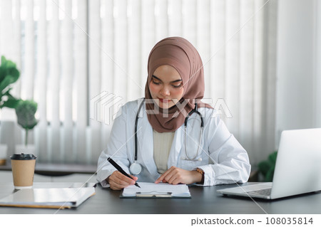 Young muslim female doctor in brown hijab headscarf working with medical papers while sitting in modern clinic office. Young muslim female doctor in brown hijab headscarf working with medical papers while sitting in modern clinic office. 108035814