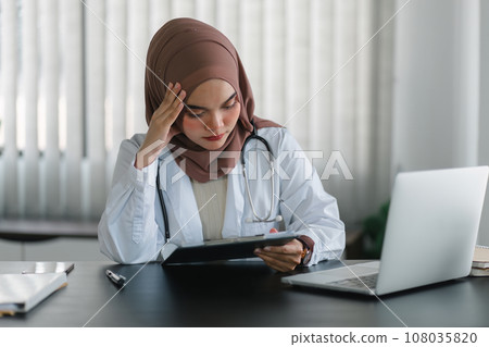 Stressed muslim female doctor in brown hijab headscarf working with medical papers looking worried, tired and overwhelmed. 108035820