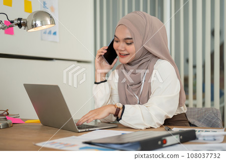 A professional Asian-Muslim businesswoman is talking on the phone with her client at her desk. 108037732