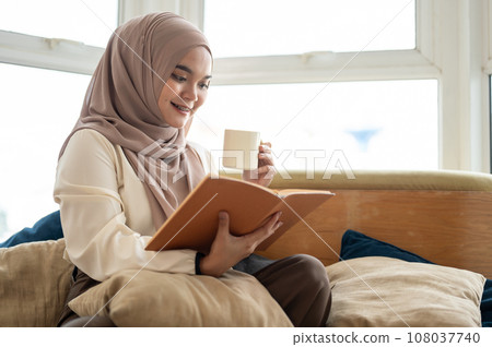 An attractive Asian-Muslim woman is enjoying her coffee while reading a book in the living room. An attractive Asian-Muslim woman is enjoying her coffee while reading a book in the living room. 108037740