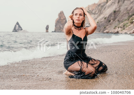 Woman summer travel sea. Happy tourist in black dress enjoy taking picture outdoors for memories. Woman traveler posing on sea beach surrounded by volcanic mountains, sharing travel adventure journey 108040706