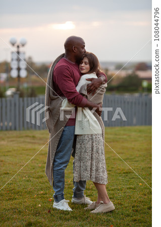 Stunning diverse couple standing in park, embraced in love, wrapped in warm blanket. Affectionate moment between African American man and Caucasian woman outdoors 108044796
