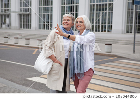 Older women with gray hair in urban landscape, smile, gaze and point off in distance. Fashionable and stylish retired female friends enjoying cityscape and adventure together Older women with gray hair in urban landscape, smile, gaze and point off in distance. Fashionable and stylish retired female friends enjoying cityscape and adventure together 108044800