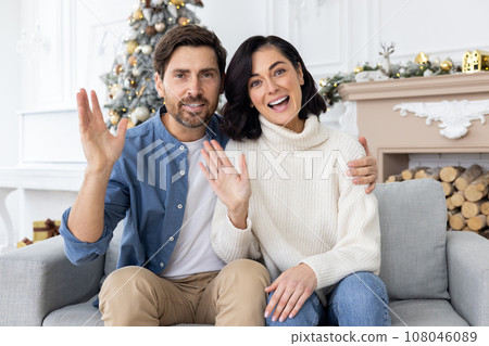 Webcam view, online video ringing, young couple man and woman looking at camera, talking with friends, congratulating on Christmas and New Year, sitting on winter day in living room, near gifts. 108046089