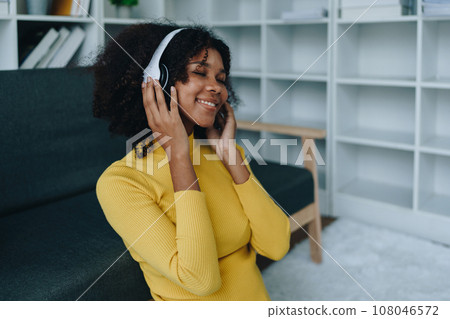 Happy young African American latina woman listening to music using headphones at home living room. relaxing, relax, relaxation concepts. Happy young African American latina woman listening to music using headphones at home living room. relaxing, relax, relaxation concepts. 108046572