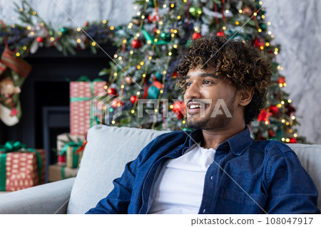 Close-up portrait of a young man for Christmas, hispanic man smiling and looking away from the window , sitting on the sofa in the living room near the Christmas tree, celebrating the new year. Close-up portrait of a young man for Christmas, hispanic man smiling and looking away from the window , sitting on the sofa in the living room near the Christmas tree, celebrating the new year. 108047917