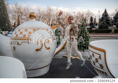 Woman Christmas Square. She stands near a large white cup, dressed in a light suit. With trees decorated with Christmas tinsel in the background 108049124