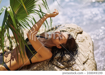 Beach Girl Model in Black Bikini With Palm Leaf on the Stone During Summer Vacation Outdoors 108049199