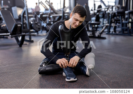 smiling young handsome sportsman with artificial leg sitting on floor at gym smiling young handsome sportsman with artificial leg sitting on floor at gym 108050124