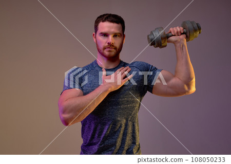 Handsome strong muscular bearded man with dumbbell touching his chest. 108050233