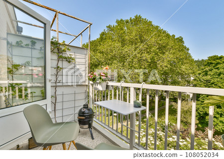 a balcony with two chairs and a grill on the left side, in front of a white fenced area 108052034