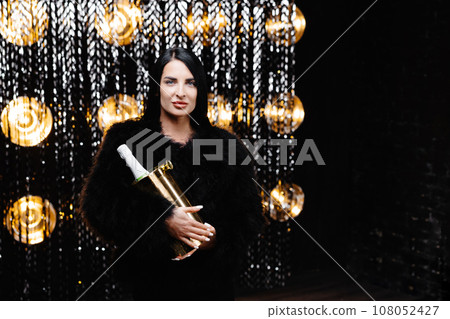 Charming woman with brunette long hair in trendy evening fur coat holding bucket with bottle with champagne on shine wall in studio Charming woman with brunette long hair in trendy evening fur coat holding bucket with bottle with champagne on shine wall in studio 108052427