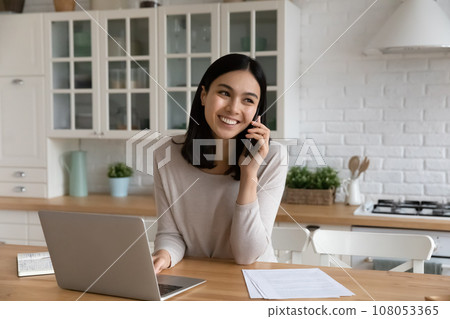 Smiling Asian woman talking on phone, using laptop in kitchen Smiling Asian woman talking on phone, using laptop in kitchen 108053365