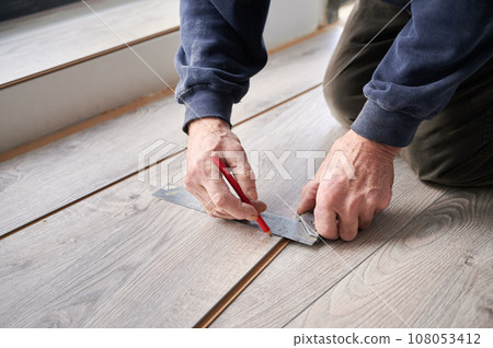 Close up of man hands using metal ruler and pencil while drawing line on laminate plank. Male construction worker measuring laminate wooden board for floor installation. Home renovation concept. Close up of man hands using metal ruler and pencil while drawing line on laminate plank. Male construction worker measuring laminate wooden board for floor installation. Home renovation concept. 108053412