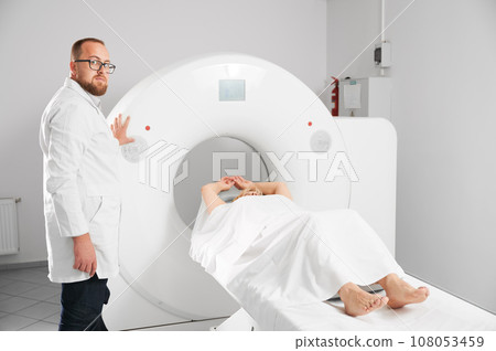 Medical computed tomography or MRI scanner. Male radiologist presses MRI button to examine female patient, looking to the camera. Medical computed tomography or MRI scanner. Male radiologist presses MRI button to examine female patient, looking to the camera. 108053459