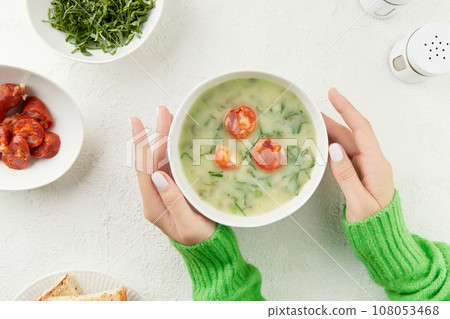 Womens hands holding bowls of Portuguese style soup Caldo Verde with bread, cabbage, and chorizo sausage on white background 108053468