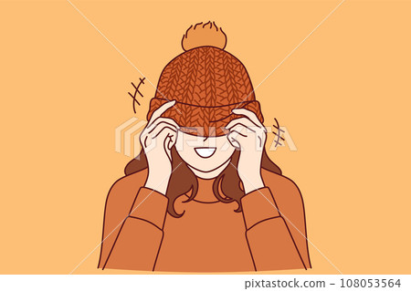 Woman covers face with knitted hat with pompom and smiles standing in warm sweater 108053564